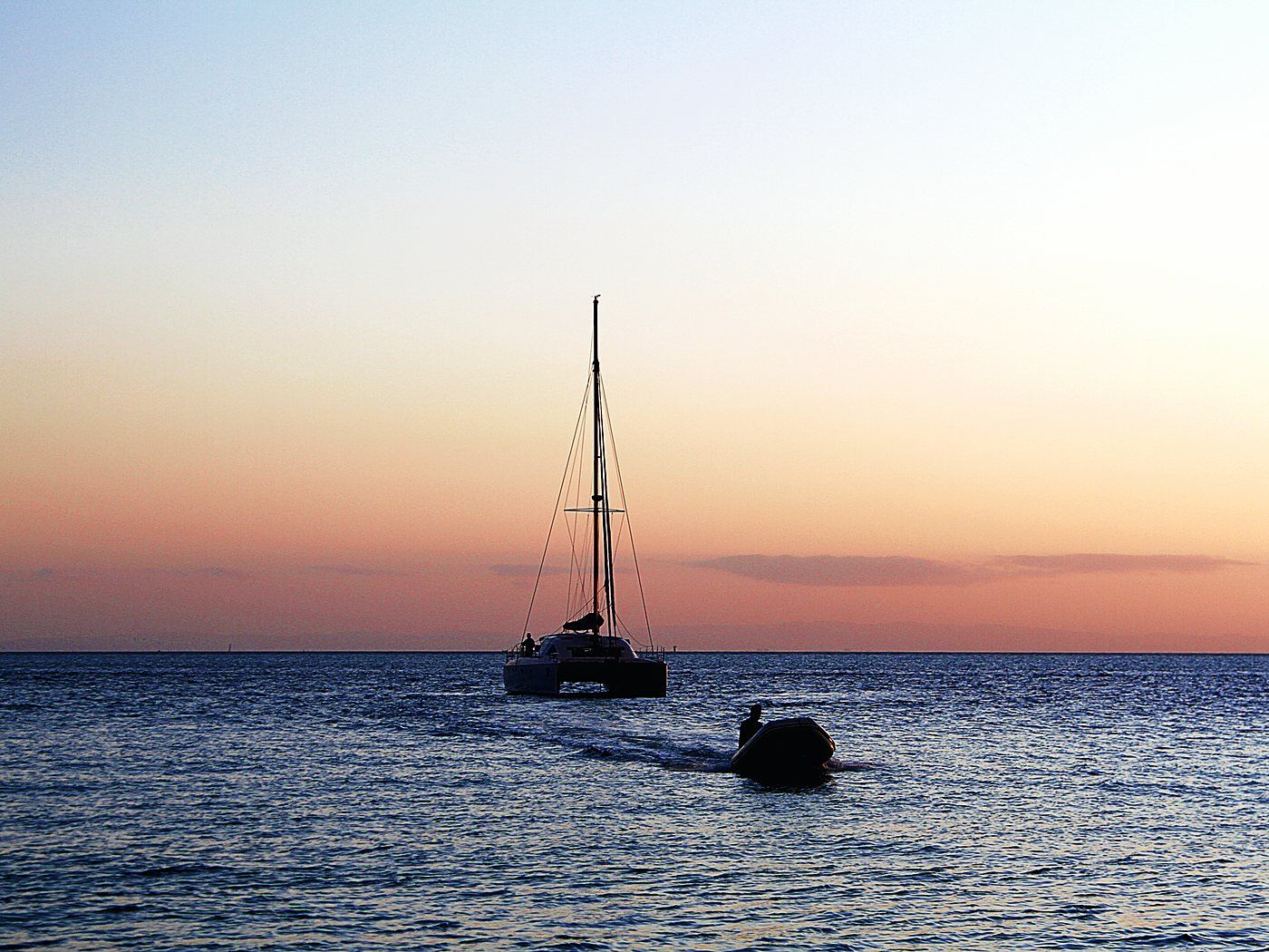 A catamaran under sail against a low sun on flat water